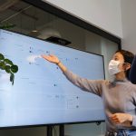 A teacher gestures to a smart board in a classroom