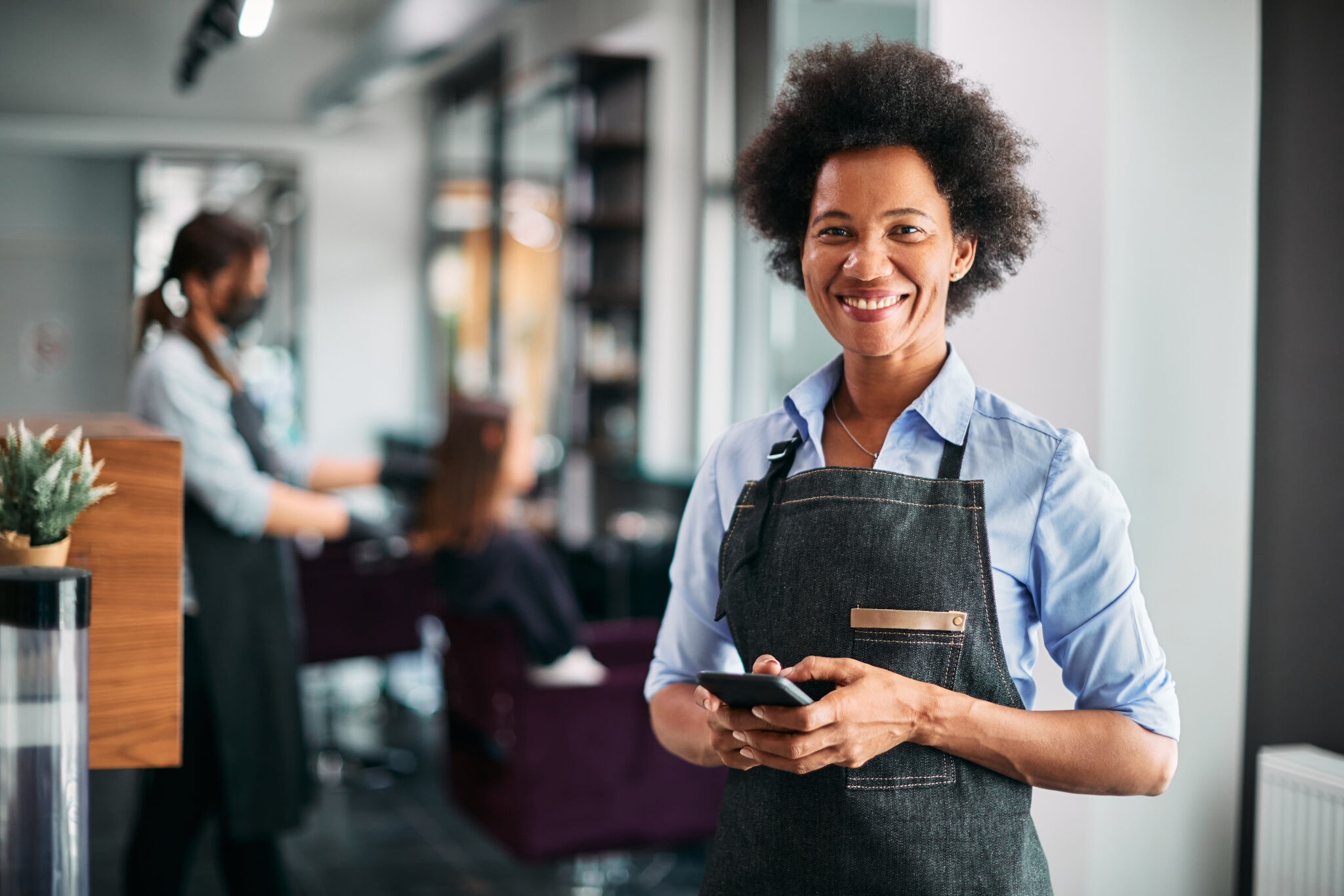 Happy African American hair salon owner text messaging on smart phone at work.